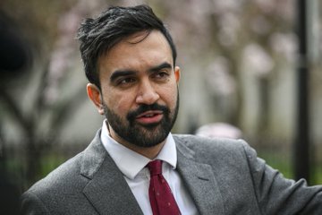 A man with dark hair and a full beard, wearing a gray suit with a white shirt and dark red tie, stands outdoors against a blurred background of trees. The image likely depicts Zohran Mamdani, who leads Polymarket's TIME Person of the Year 2026 odds at 22%, ahead of Trump (21%), Pope Leo XIV (12%), and Netanyahu (11%).