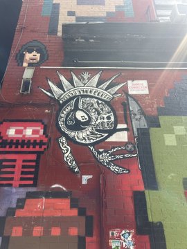 A pixel-art style mural painted on a red brick wall, featuring a crowned skeleton character holding a scythe, with green and grey blocky elements.