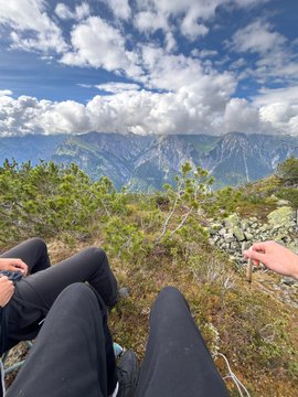 The image shows two people sitting next to each other with their legs crossed while looking out at a beautiful mountain range or valley below them. The scene appears to be taken from an elevated position, possibly from a chair or a hilltop, which provides a unique perspective of the landscape. The photo has been edited to include text that reads "Oh, how good it feels to be a $neet". This phrase is likely meant as a playful and humorous response to the image's title, suggesting that the two people are enjoying their time together in this picturesque setting.
