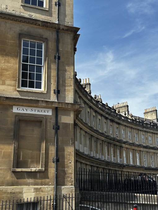 A curved row of Georgian-style stone buildings along Gay Street, with a street sign visible on the corner building under a partly cloudy sky.