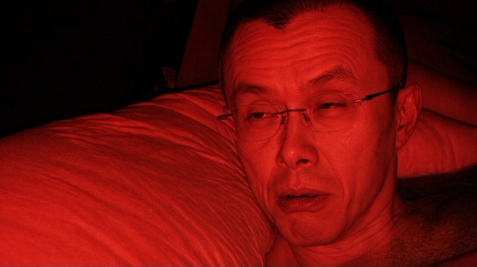 A man's face illuminated in red light while lying on a white pillow.