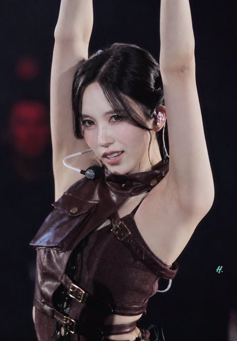 A female performer with dark hair wears a brown leather harness outfit and headset microphone while performing on stage with her arms raised.