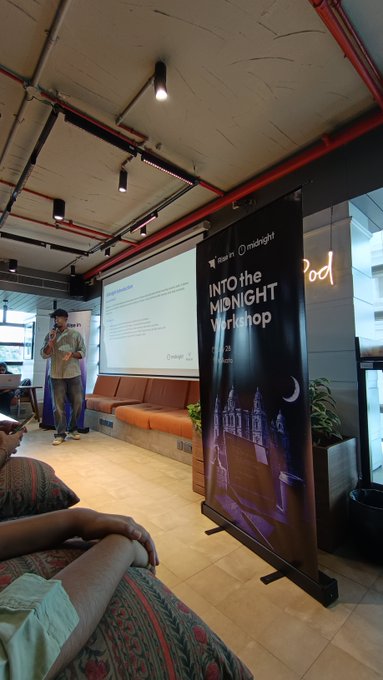 A presentation room with industrial ceiling featuring a banner reading 'INTO THE MIDNIGHT Workshop' sponsored by InMidnight, with a speaker presenting to a small audience.
