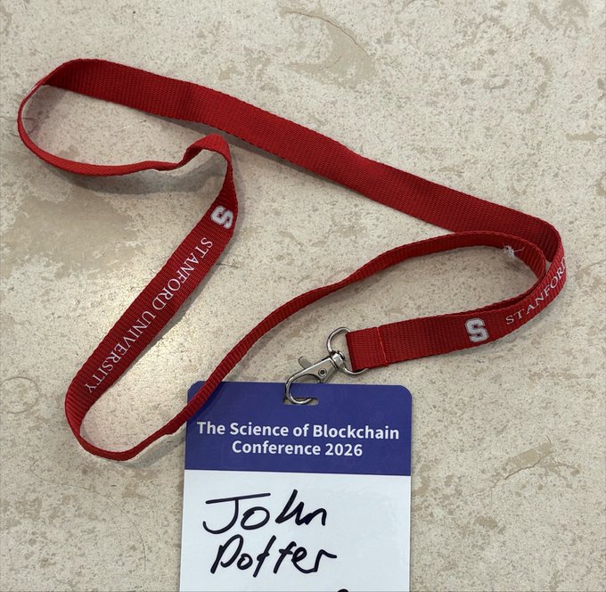 Red Stanford University lanyard with a conference badge reading "The Science of Blockchain Conference 2026" and the name "John Poffe" handwritten on it.