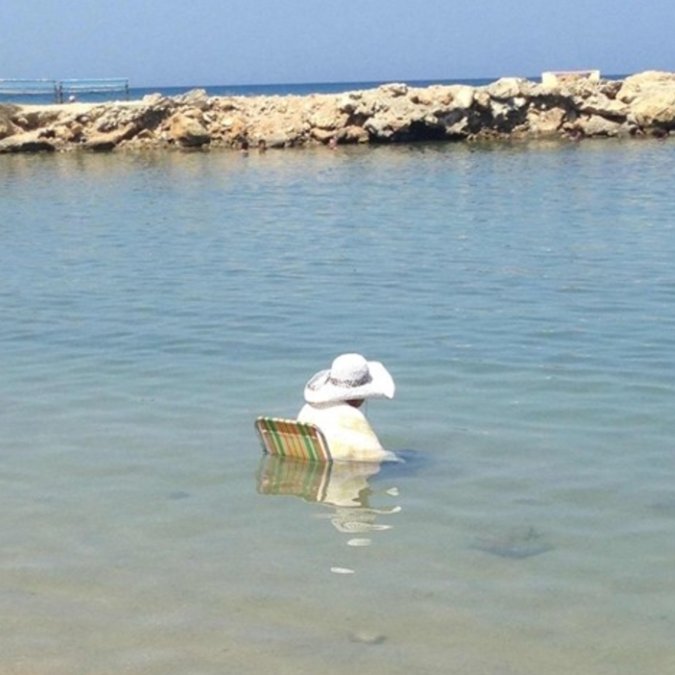 The image shows a person sitting in shallow water, wearing a white hat and holding onto a floating object or board. The water appears to be calm with no visible waves or ripples. The surrounding area includes rocks and a sandy beach, suggesting that this scene takes place near a body of water like an ocean or sea. The post reads "hyperliquid", which could possibly be a reference to the person's attire (white hat) or the water's characteristics (calm).