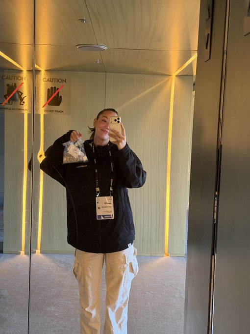 A woman takes a mirror selfie in an elevator holding a food container, with 'CAUTION DO NOT TOUCH' signs visible on the mirrored walls behind her.
