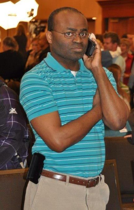 A serious-looking man in a teal striped polo shirt and khaki pants is talking on a mobile phone, with a visible black holstered handgun at his waist, suggesting the FBI agent referenced in the caption responding to a bomb threat at the conference.