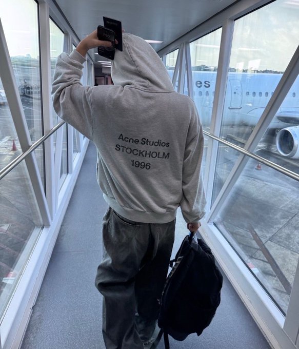 A person walks down an airport jet bridge wearing a beige "Acne Studios Stockholm 1996" hoodie and carrying a black backpack, with a plane visible through the window.