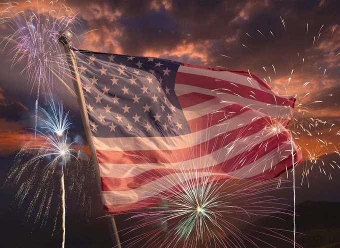 An American flag waves dramatically against a sunset sky filled with exploding fireworks in red, white, and blue colors.