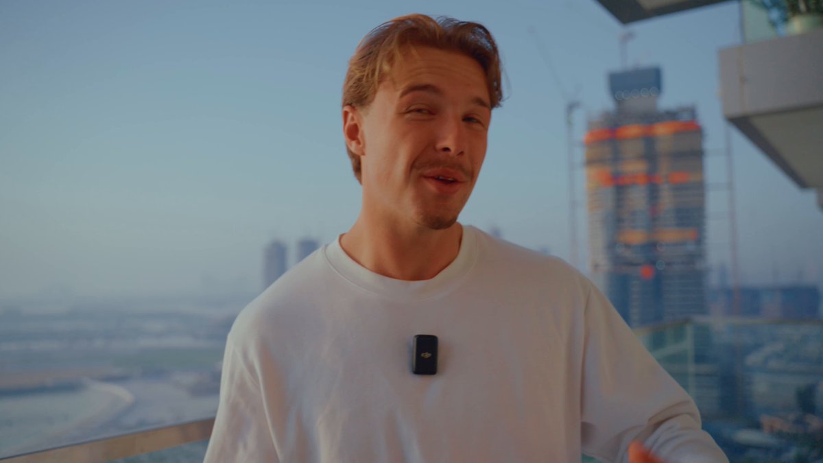 The image features a man wearing a white shirt with a black object attached to his neck. The man appears to be standing near a balcony or deck, possibly at an apartment complex. In the background, there are buildings that could potentially be part of a cityscape. The main focus of the image is on the man and the object he's wearing.