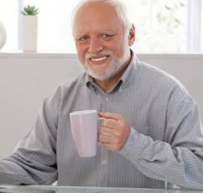 A smiling older man with a white beard holds a white mug, wearing a light blue checkered button-down shirt in a bright indoor setting.