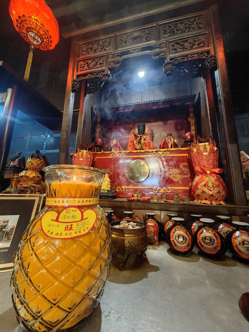 A large golden pineapple-shaped jar of "Tuak 胜男" rice wine sits before a traditional Chinese temple altar, where deity statues are surrounded by smoke from burning incense and rows of smaller ceremonial jars. The image captures a moment of prayer and offering, likely at a temple in Borneo (Sarawak), fitting the poster's hopeful message seeking blessings of good luck, health, and energy for the days ahead.