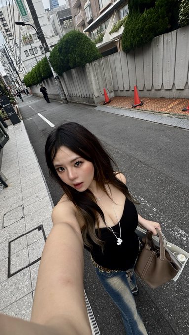 A selfie of a woman with long brown hair wearing a black tank top, blue jeans, and a heart pendant necklace, holding a beige handbag on a city street.
