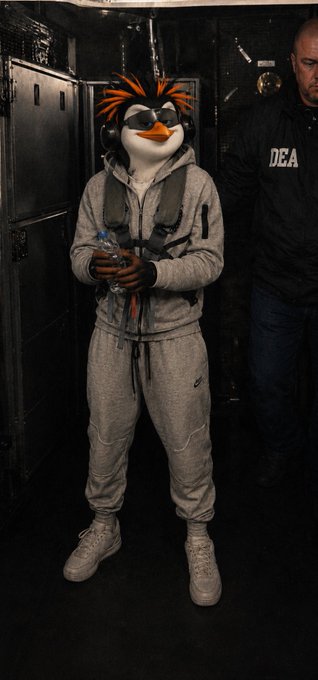 A person in a gray Nike tracksuit wearing a penguin mascot head with orange and black hair and headphones stands next to a man in a "DEA" jacket. The surreal, meme-like image fits the casual, quirky tone of a "Gm" (good morning) crypto greeting.