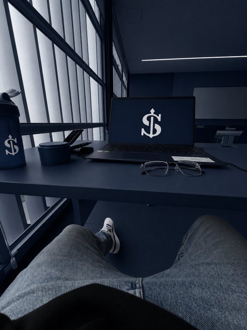 A first-person view of someone sitting at a desk with a laptop showing a dollar sign ($) logo on the screen, alongside a matching shaker bottle, eyeglasses, and a dark blue minimalist workspace.