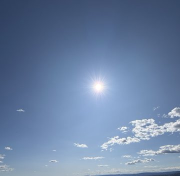 A bright photograph of the sun shining in a clear blue sky with small scattered clouds.