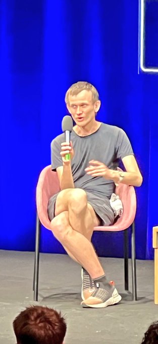 A man in a gray t-shirt and shorts sitting on a pink chair on a stage, holding a microphone and speaking to an audience against a blue curtain backdrop.