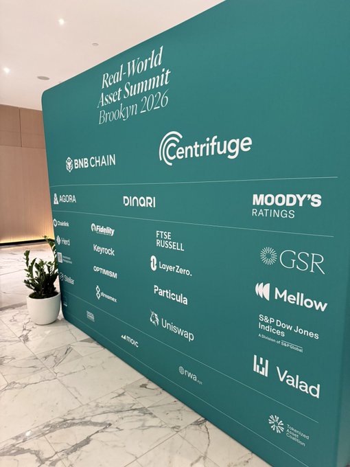 A green sponsor wall displays the "Real-World Asset Summit Brooklyn 2026" title, featuring logos from partners like BNB Chain, Centrifuge, Moody's Ratings, Chainlink, and Uniswap, reflecting the event's strong industry turnout in line with the bullish RWA sentiment.