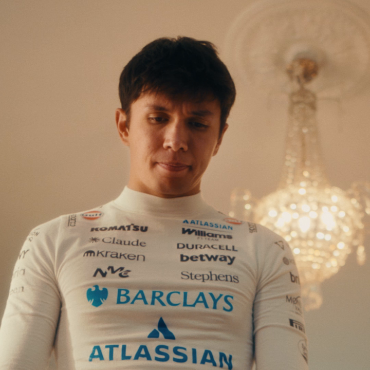 A young man with dark hair stands in an elegant setting with a chandelier in the background, wearing a white Williams Racing racing suit featuring sponsor logos including Barclays, Atlassian, Komatsu, Kraken, Claude, Duracell, Betway, and Gulf. The image ties to the post by visually pairing the Williams F1 driver with the sponsor brands prominently displayed on his race suit, highlighting the connection between the team and its partners promoted by the cryptocurrency and stocks app.