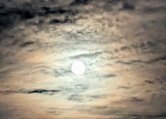 A photograph of a bright full moon glowing through a layer of wispy, textured clouds in a pale sky.