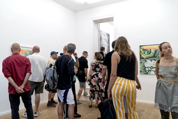 A diverse group of gallery visitors gathers inside a bright, white-walled exhibition space, observing colorful artworks displayed on the walls during what appears to be the opening of the "STILL WET" exhibition in Berlin.