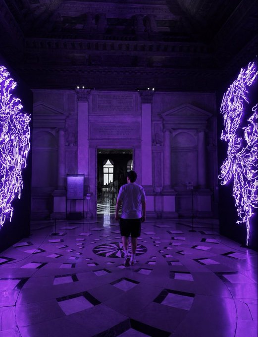 The image shows a person walking through an art gallery filled with purple neon lights. The person appears to be admiring the artwork or simply passing by. The gallery features two large pieces of art, one located near the center of the room and another towards the right side. The scene is illuminated by the vibrant purple lights, creating a unique atmosphere in the space.