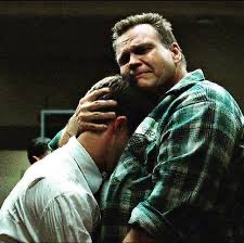 Two men embrace in a hug, one in a white shirt and the other in a green plaid shirt, in what appears to be an emotional scene.