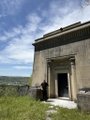 The image features an old stone building situated at the top of a hill or mountain. The building has a small door located near its entrance, which appears to be open. In front of the building, there are two barrels placed next to each other on the ground. The scene is captured from a low angle, giving prominence to the building and the barrel arrangement.