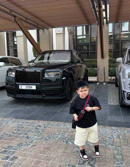 A young boy wearing a black shirt, beige shorts, and a black cap posing with a peace sign in front of a black Rolls-Royce with Dubai license plate D 57594.
