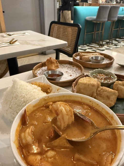 A meal at a restaurant featuring a bowl of curry with shrimp, white rice, spring rolls, fried tofu, and dipping sauces on a marble table.