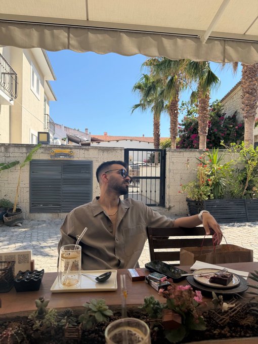 A man wearing sunglasses and an olive shirt relaxes at an outdoor cafe table with a drink and dessert, with palm trees visible in the background.