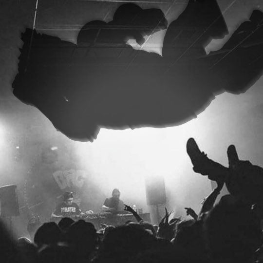 A large crowd of people gathers at a concert, with the stage illuminated by bright lights and a DJ performing on a drum set.