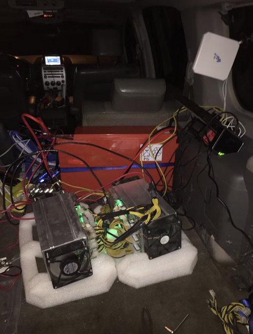 A makeshift mining rig consisting of two Antminer S9 Bitcoin miners (each with cooling fans) sits on foam padding in the back of a vehicle, wired with tangled yellow, red, and black cables running to power supplies. This raw, early setup—delivering approximately 20 TH/s directly from the car's alternator—represents the humble, mobile origins of what would become a full-scale mining operation.