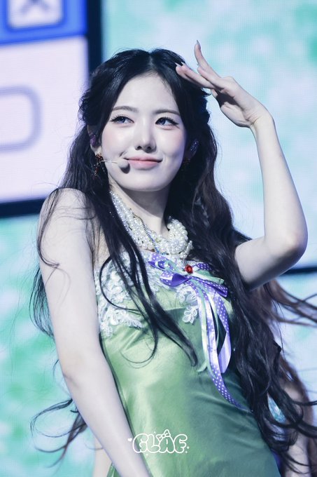 A young woman with long dark hair performs on stage wearing a green and white outfit with a large pearl necklace and ribbons, making a playful gesture near her head.