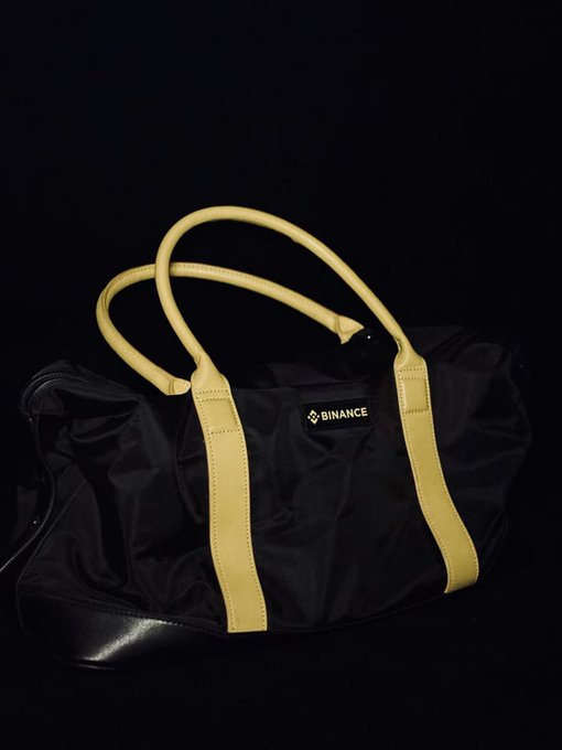 A black duffel bag with yellow leather straps and handles features a 'BINANCE' logo patch on the front, displayed against a dark background.
