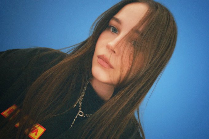 A portrait of a young woman with long brown hair and striking blue eyes, posed against a solid blue background.