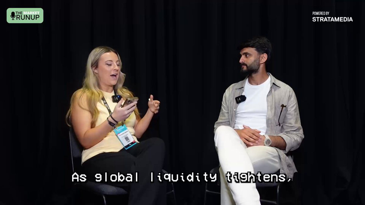 The image shows a man and woman sitting next to each other, with the woman holding a cell phone while talking to the man. The background of the image features a black curtain, which appears to be part of an event or presentation setting. There is also text in the image that reads "As global liquidity fights", possibly referring to a discussion about the current state of the cryptocurrency market.
