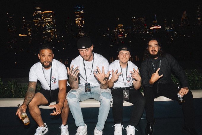 Four men posing together at night with a city skyline behind them, three wearing matching white t-shirts and black caps while making hand gestures, while the fourth on the right wears all black.