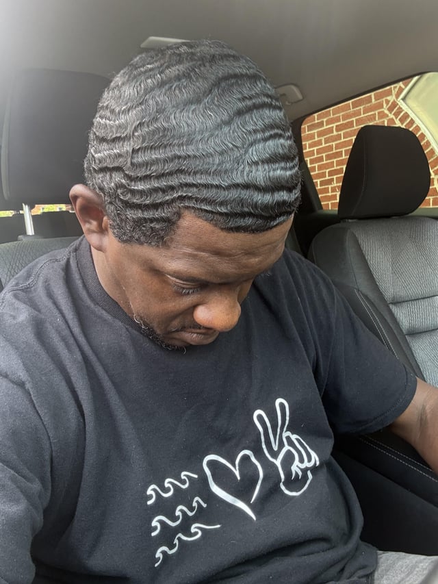 A man sits in a car wearing a black t-shirt featuring a "love" graphic with a peace sign and wave, complementing the "✌🏾❤️🌊" emojis in the post, while his freshly styled waves match the Hedera-themed reference.