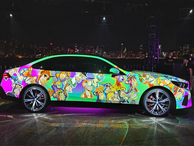 A side view of a BMW car featuring vibrant, colorful artwork with cartoon characters projected or wrapped onto its exterior at night, with city lights in the background.