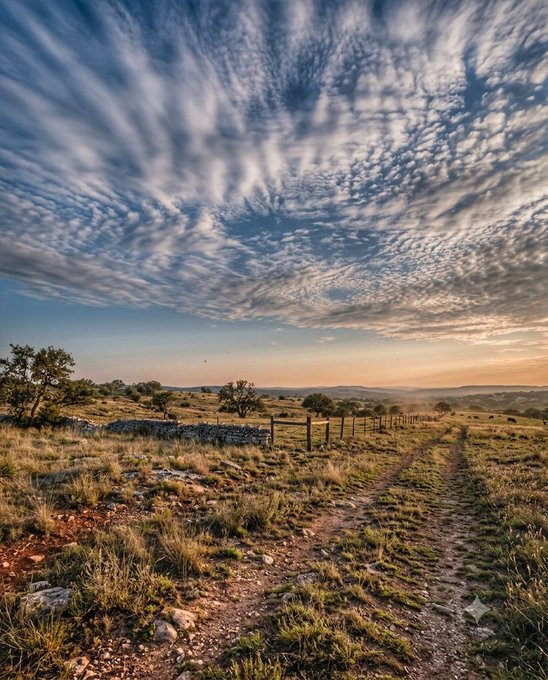 A vast, rolling grassland under a dramatic sky filled with scattered altocumulus clouds at sunset, with a dirt trail and stone wall leading into the distance. The image's serene, expansive landscape evokes the hopeful "keep going" sentiment of the post, visually reinforcing the encouragement to buy more $XRP and support $XTZ artists.