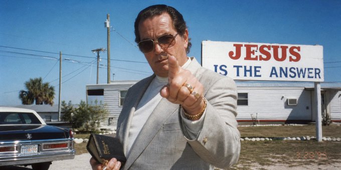 A man in a tan suit and aviator sunglasses points at the camera while holding a Holy Bible, standing in front of a large sign that reads 'JESUS IS THE ANSWER' with a building, palm trees, and a parked car in the background.
