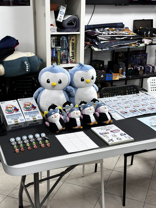 A merchandise table displaying various plush toys, including penguin and sheep characters, small character figurines, and photo cards on a folding table.