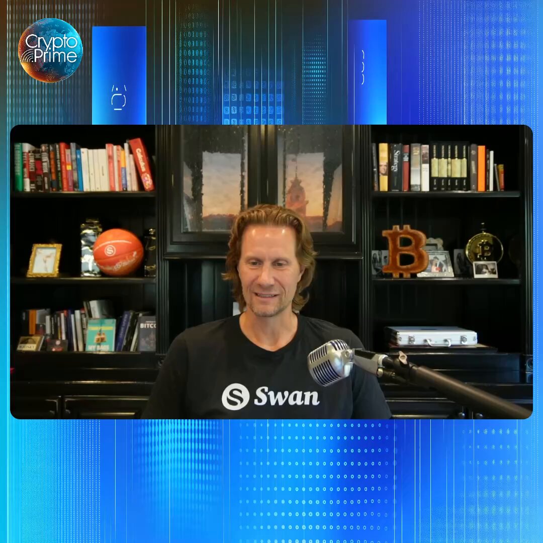 The image features a man wearing a black shirt with "Swan" written across his chest, sitting behind a microphone at a desk or table. The background of the image shows a blue wall with various books scattered around, suggesting that he might be in a library or a workspace. There is also an orange sports ball on the left side of the image.