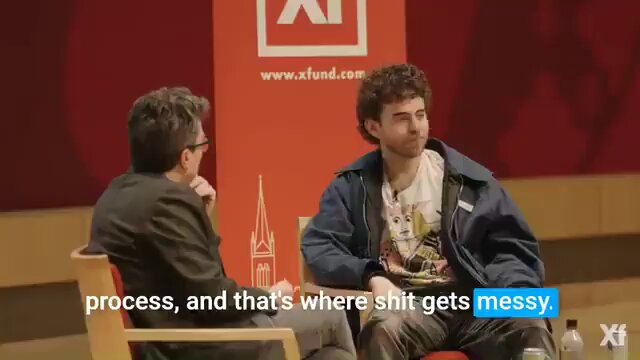 The image shows two men sitting next to each other, having a conversation or talking to someone who appears to be recording them. There are also some words related to the stock market visible in the scene. The primary focus of the image is likely on these two individuals and their interaction with the recorder.