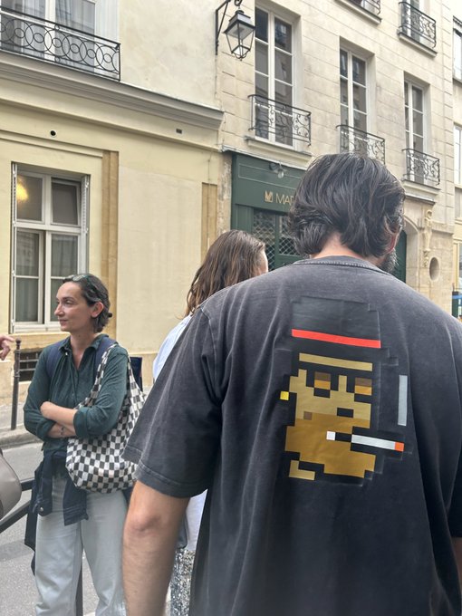 A street scene in Paris where a man wears a black t-shirt featuring a pixelated art graphic on the back, with a checkered bag visible on another person nearby.