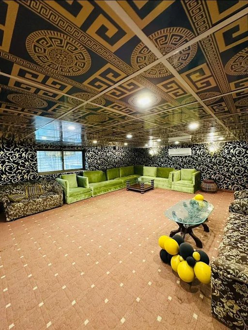 An ornate interior room features a highly decorated ceiling with gold Greek key patterns, dark floral wallpaper, green velvet sectional sofas, and a cluster of yellow and black balloons.