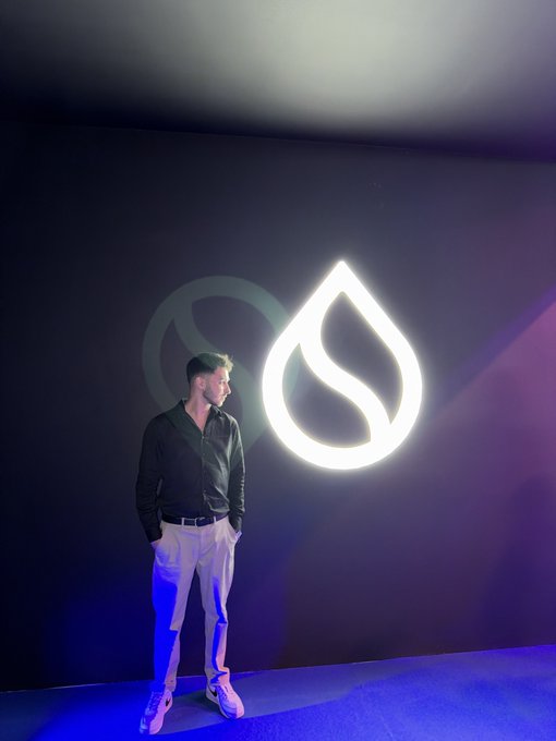 A man in a black shirt and beige pants stands against a dark wall featuring a large illuminated teardrop or flame-shaped logo.