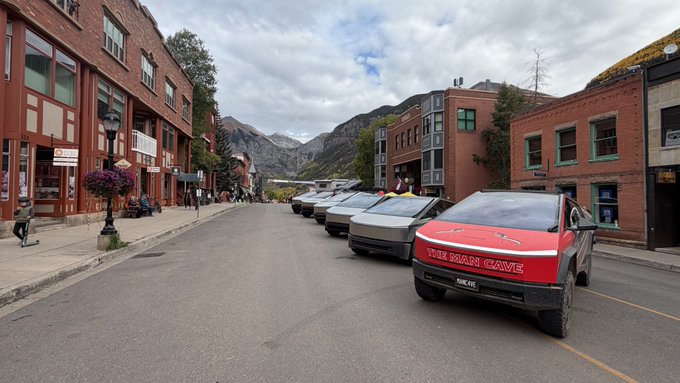 A lineup of Tesla Cybertrucks labeled "THE BRANT CARVE" are parked along a quaint mountain town street, building anticipation for an exciting upcoming reveal or showcase.
