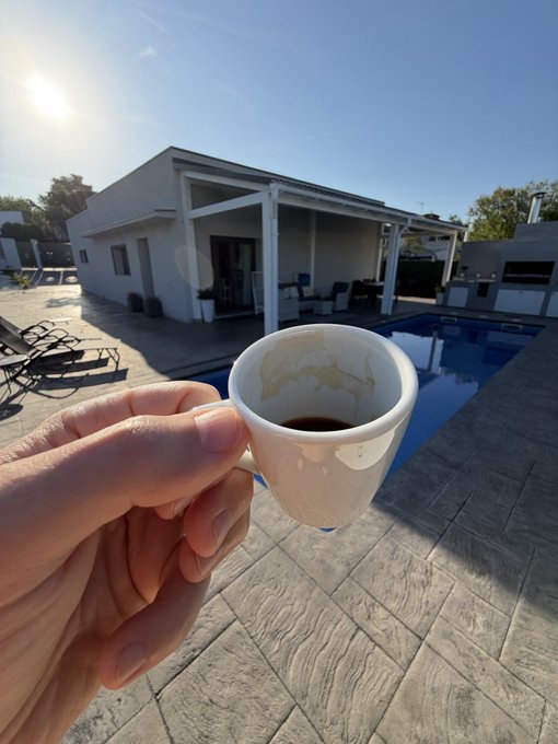 A hand holds a white mug of coffee in the foreground, with a modern house featuring a covered patio and a swimming pool visible in the background on a sunny day.